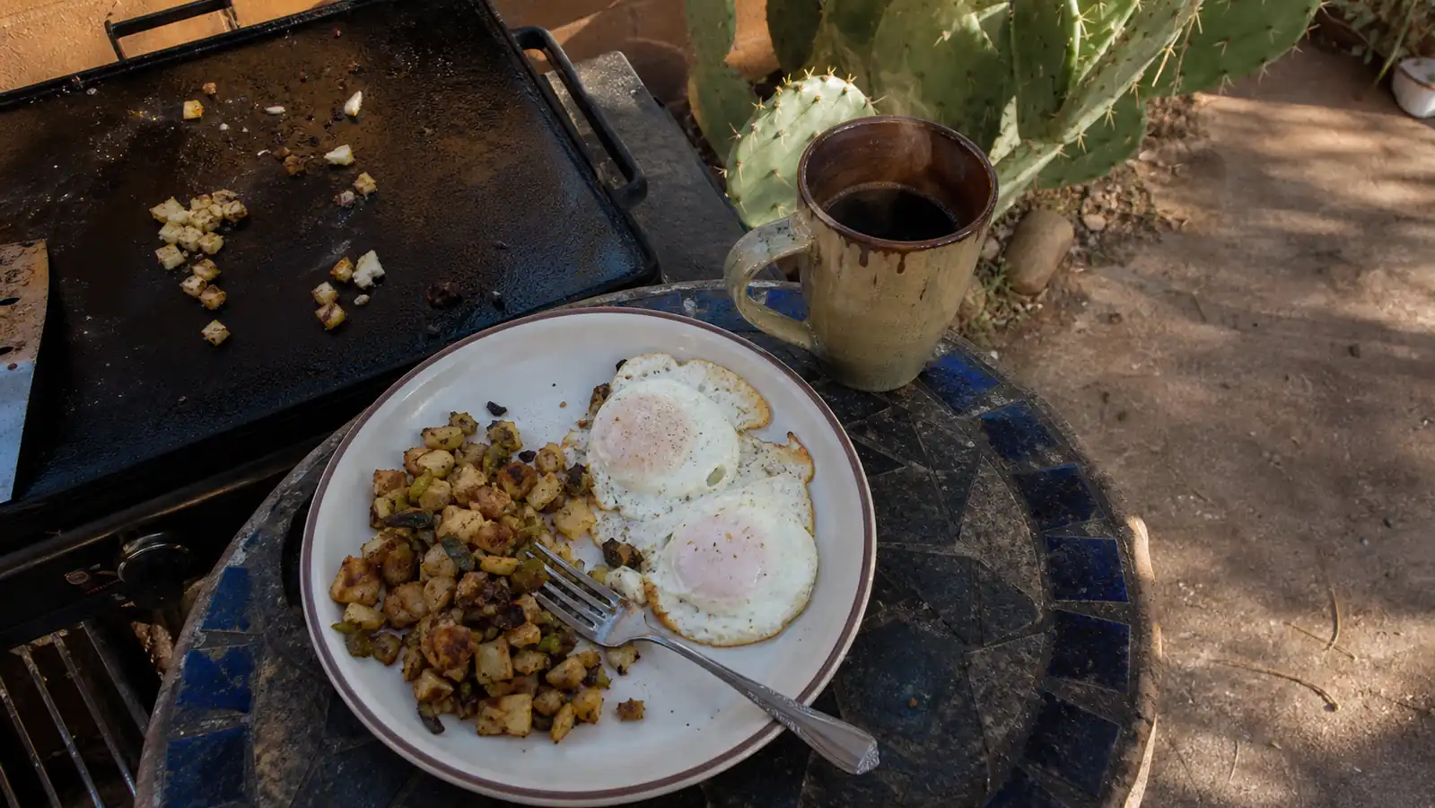 A plate of eggs and green chile hash browns cooked on a cast iron griddle set outdoors on a patio table next to a steaming coffee mug