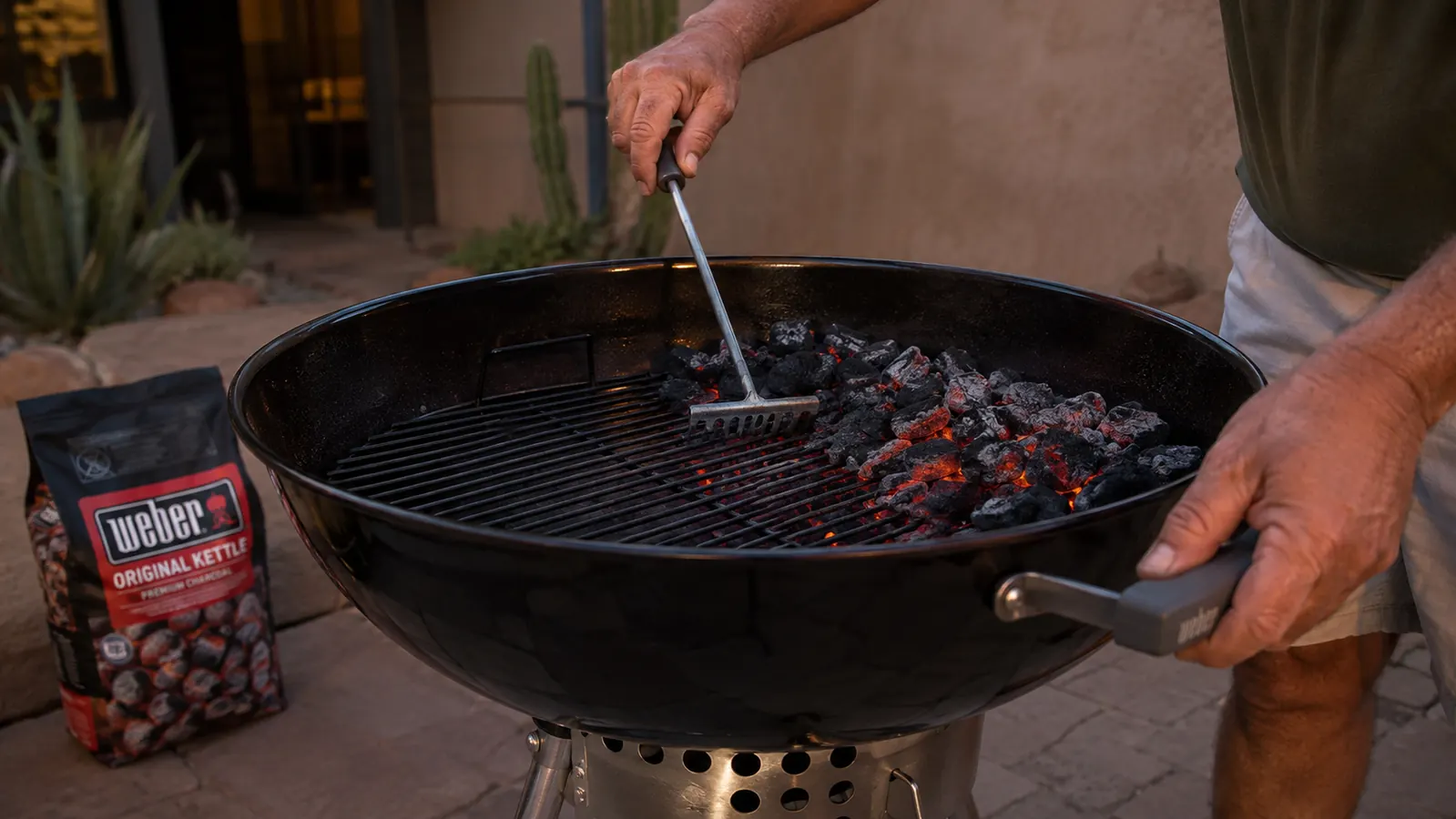 A man's hands banking glowing coals to one side of an open kettle grill using a metal rake