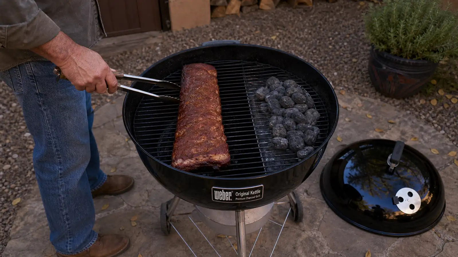 A kettle grill lid resting off to the side while a rack of ribs cooks over indirect coals on a fall evening