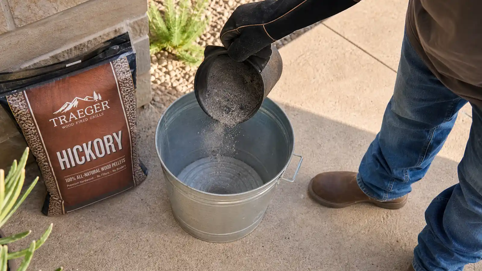 A gloved hand tipping a small amount of fine gray ash from a smoker firepot into a metal bucket