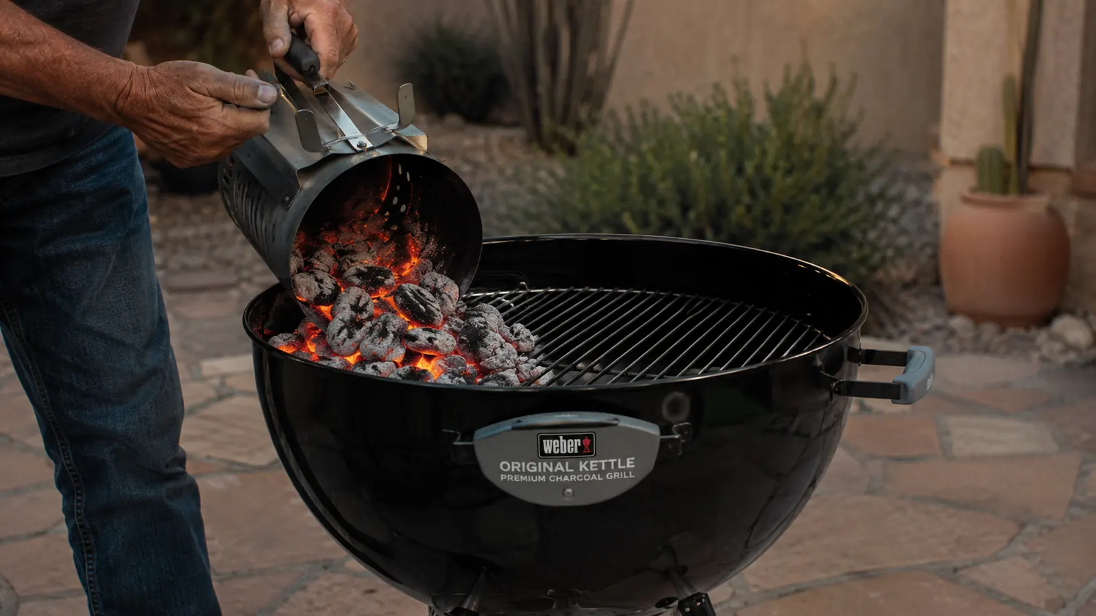 A man's hands pouring glowing coals from a chimney starter into one side of an open kettle grill to bank a two-zone fire