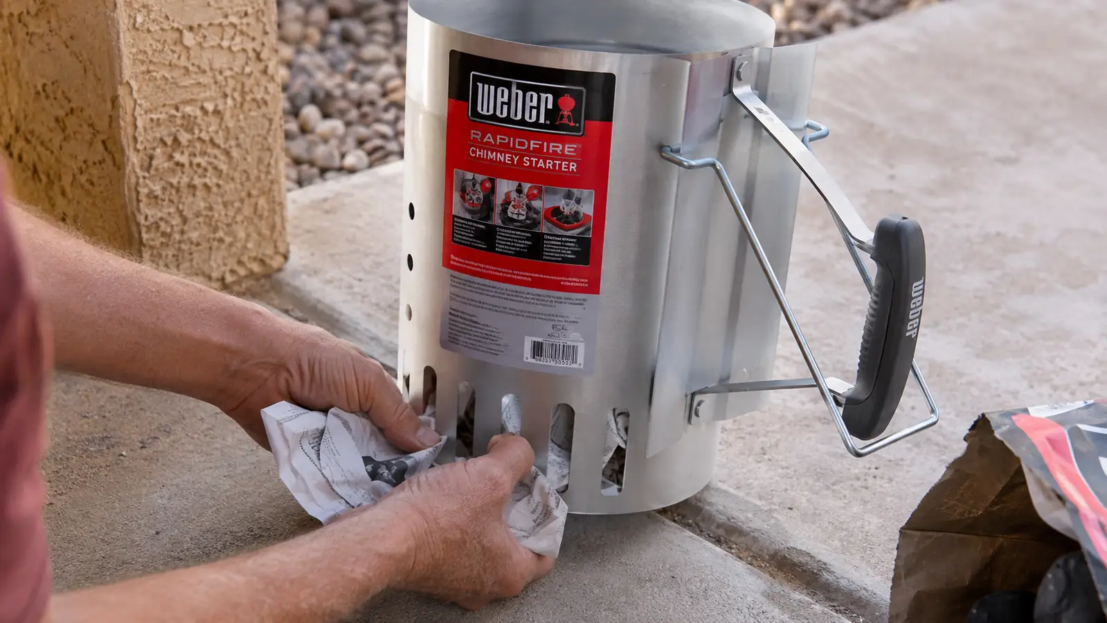 Hands tucking crumpled newspaper into the bottom vent holes of a chimney starter set on a concrete patio