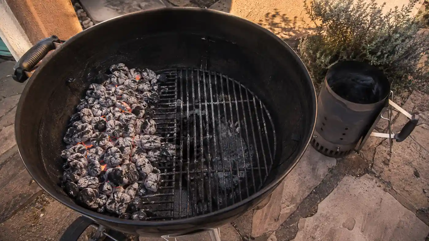 Two-zone charcoal fire banked to one side inside an open kettle grill with the empty chimney starter cooling on the patio nearby
