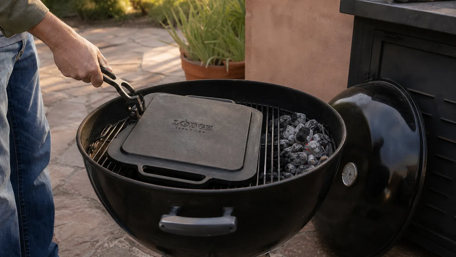 A cast iron griddle set upside down on the open grates of a kettle grill with the lid propped nearby during a heating cycle