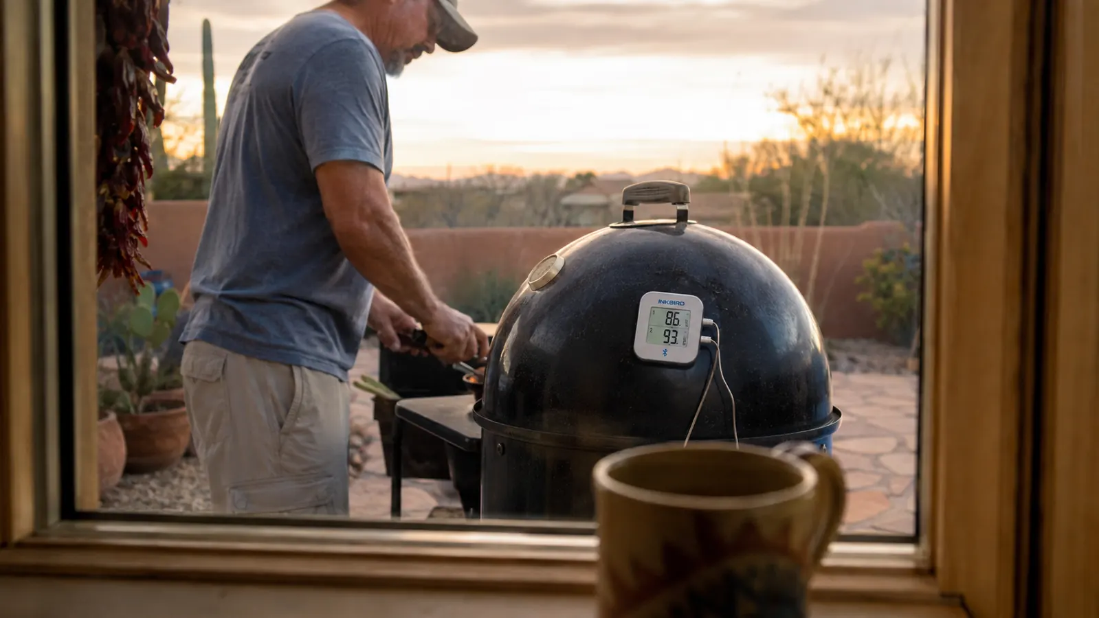A small digital thermometer base station mounted with a magnet to the curved hood of a kettle smoker, seen through a kitchen window