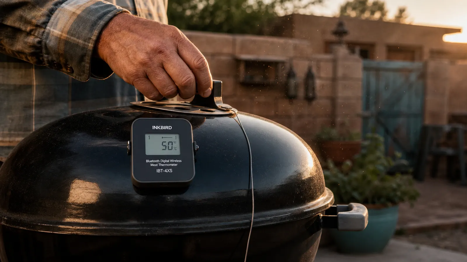 A base station thermometer mounted with a magnet to the curved hood of a kettle smoker with a probe wire running to the grate