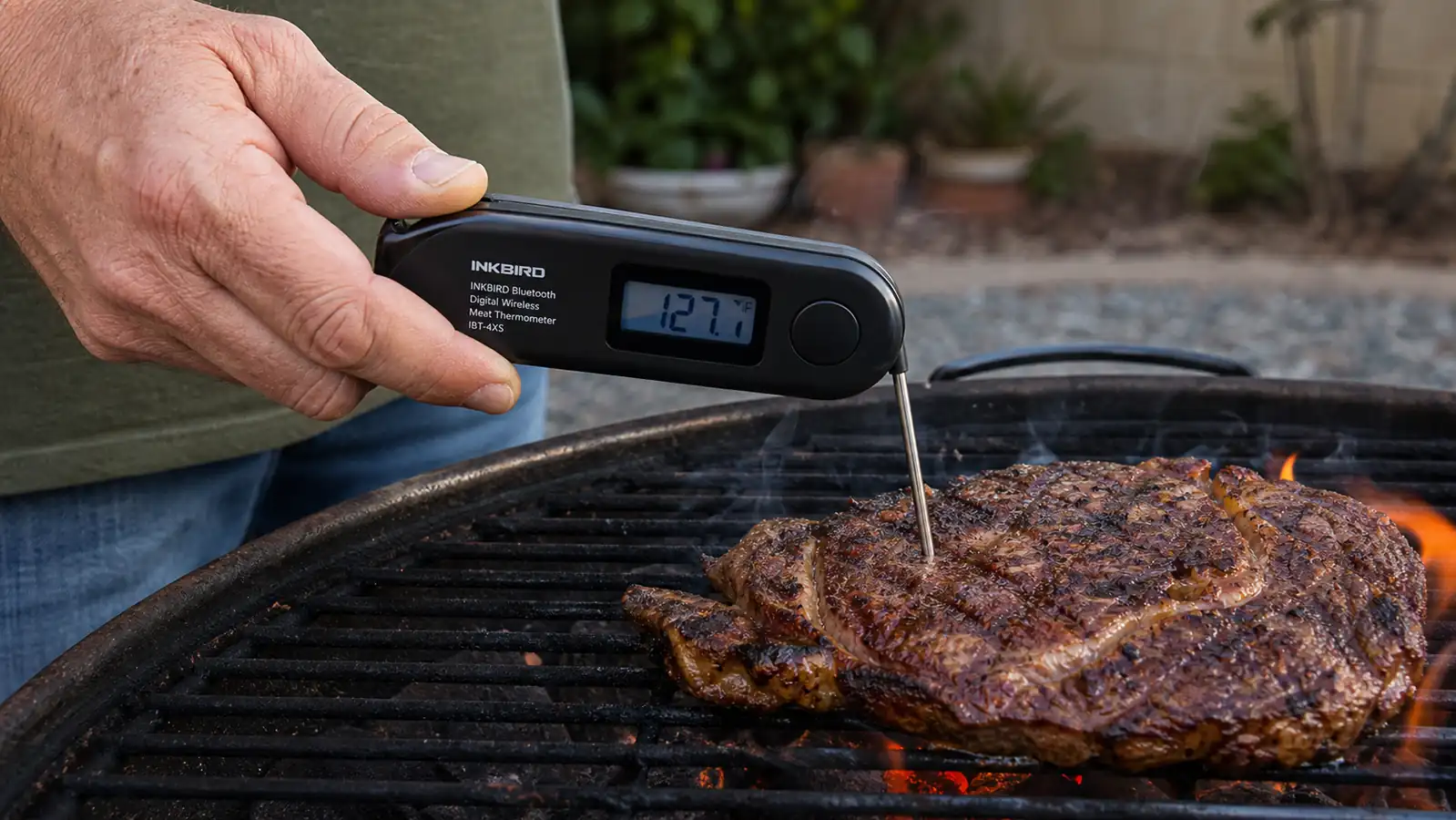 A hand holding a folding instant-read thermometer probe against the surface of a seared steak on a hot grate