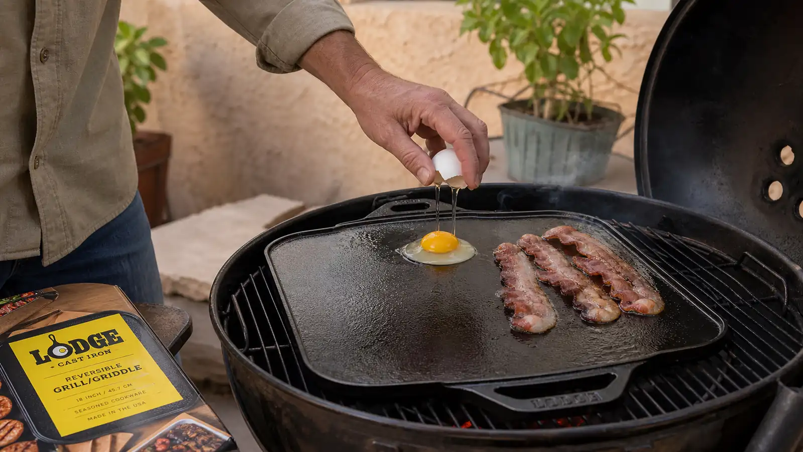 A hand cracking eggs onto one half of a hot cast iron griddle while bacon strips cook on the other half outside