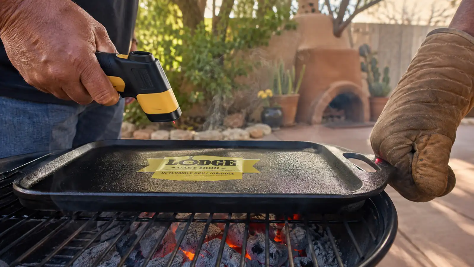A hand in an oven mitt reaching toward a griddle handle while an infrared thermometer is pointed at it over glowing coals
