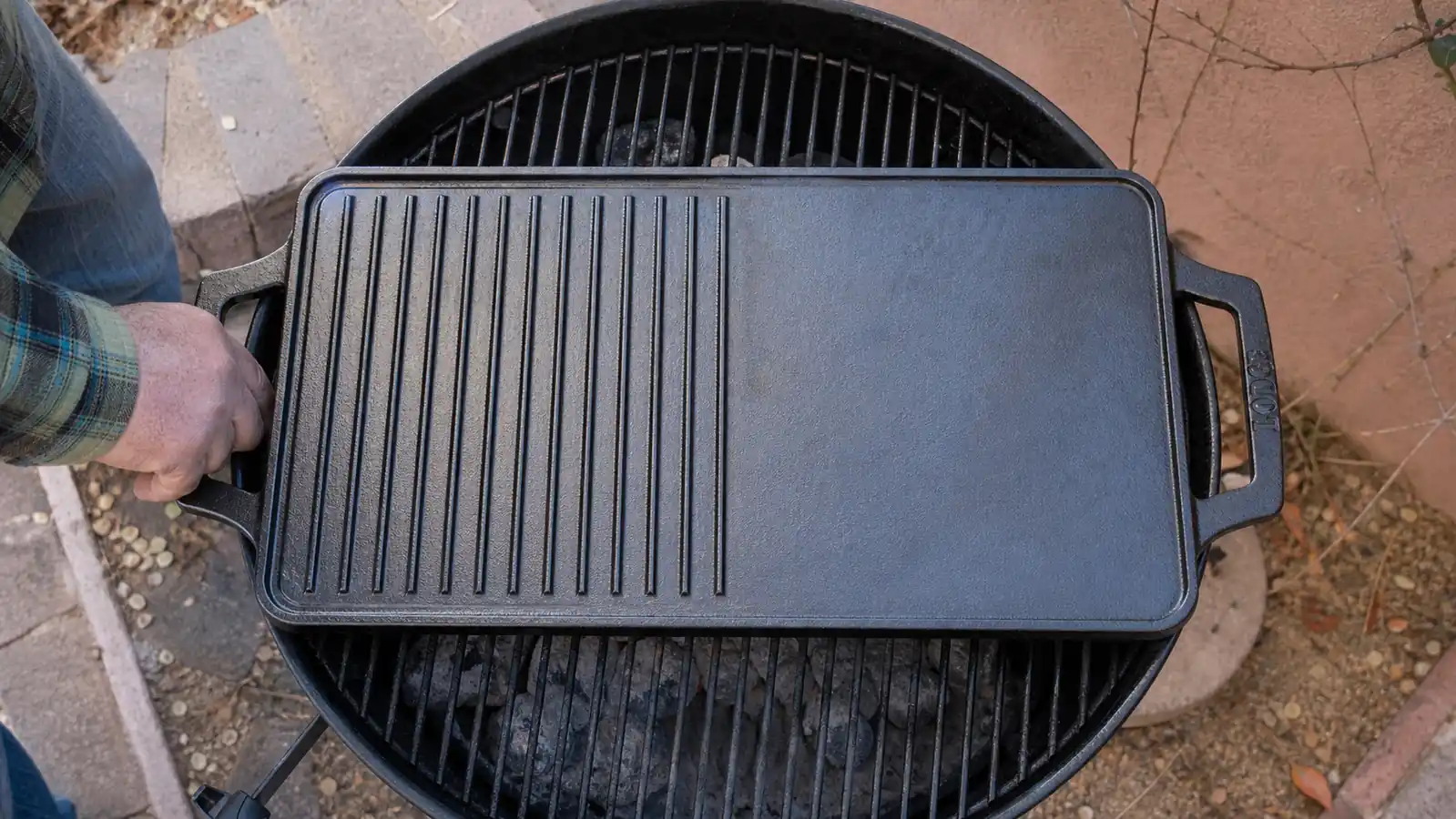 Overhead view of a rectangular cast iron griddle set across the round grate of a kettle grill with its corners overhanging the edge