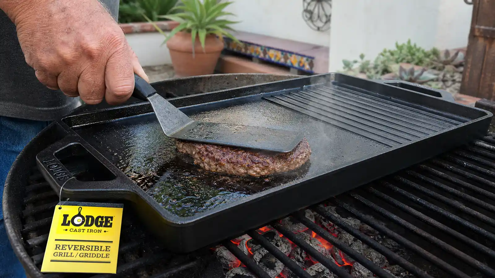 A hand pressing a metal spatula down on a smash burger patty on the flat side of a cast iron griddle over open coals