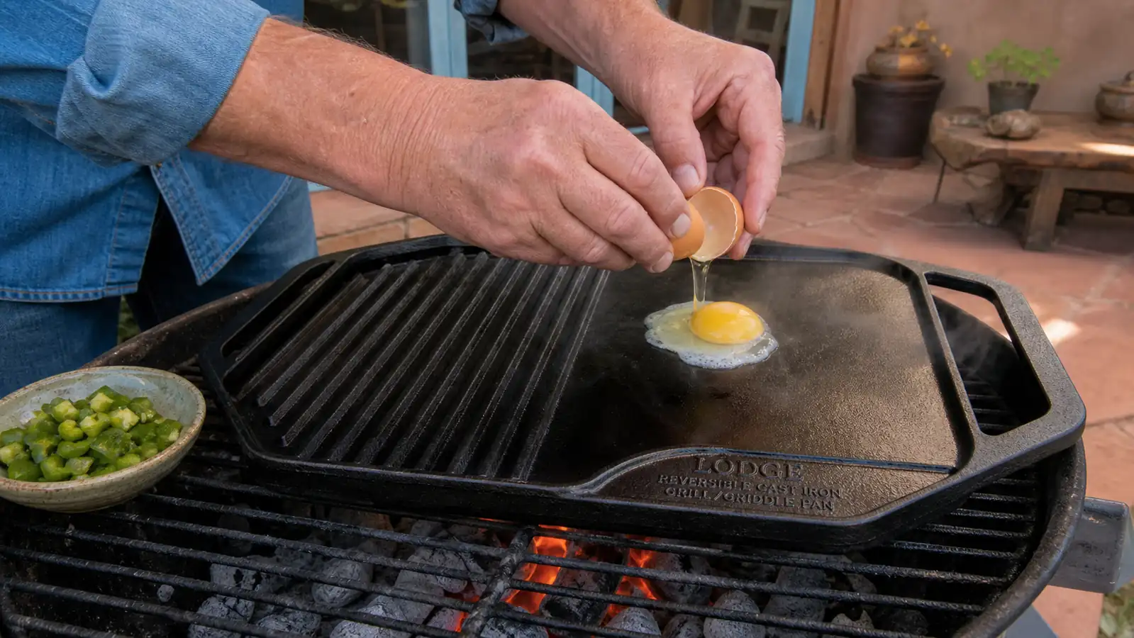 A hand cracking an egg onto the flat side of a cast iron griddle sitting over glowing coals