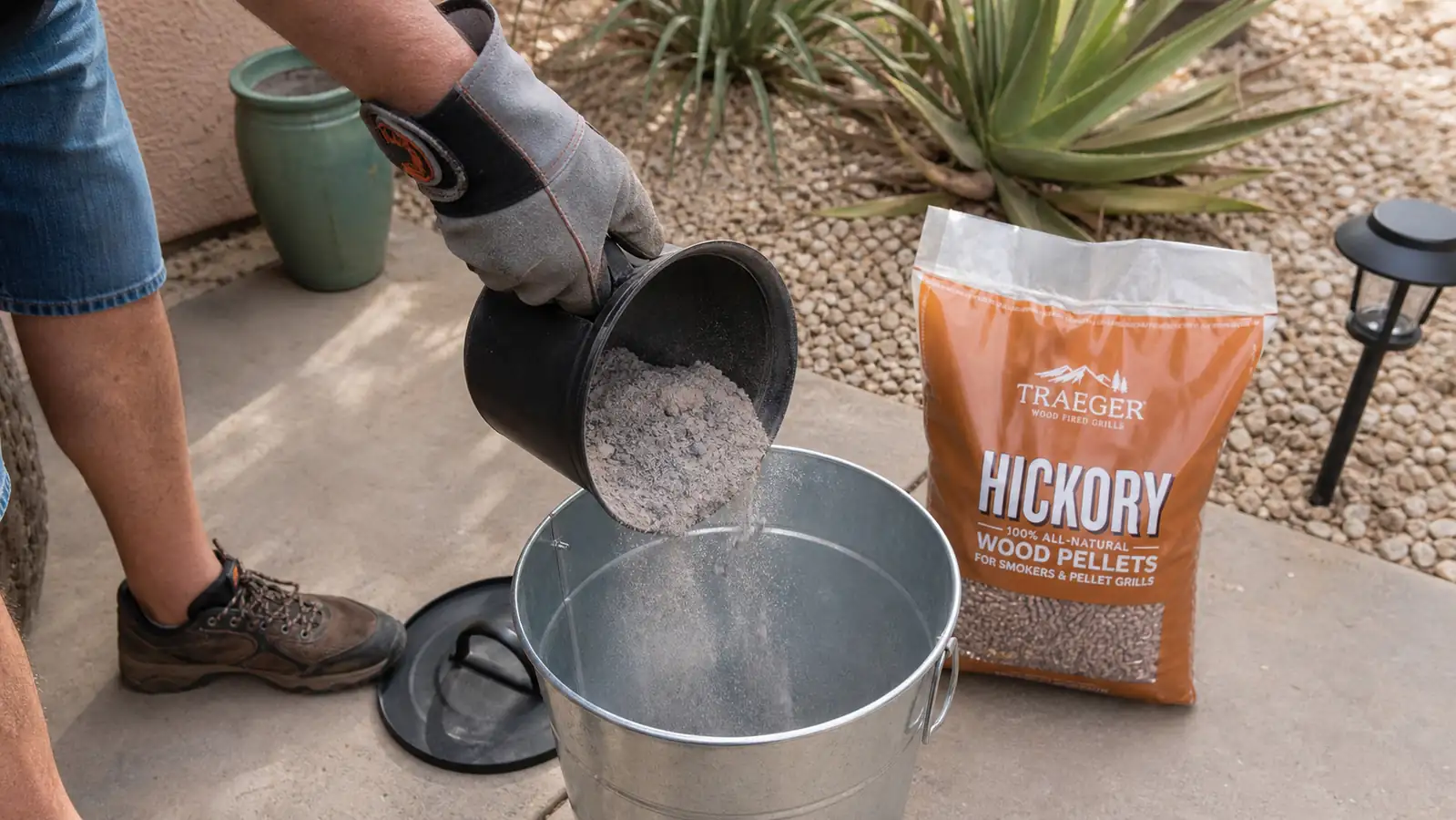 A gloved hand emptying a small pile of fine gray ash from a smoker firepot into a metal bucket
