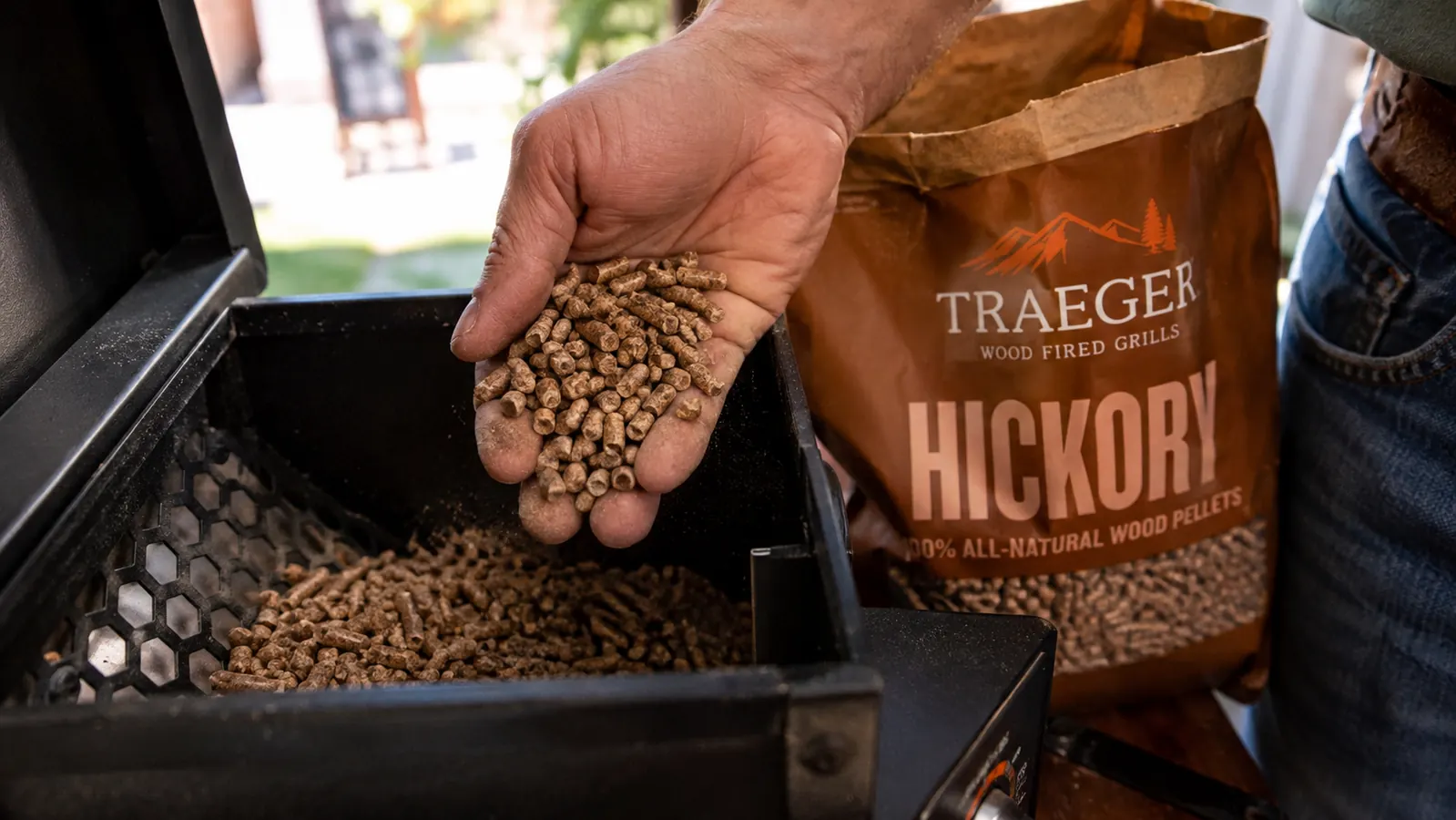 A weathered hand scooping golden-brown wood pellets from an open bag into a smoker hopper