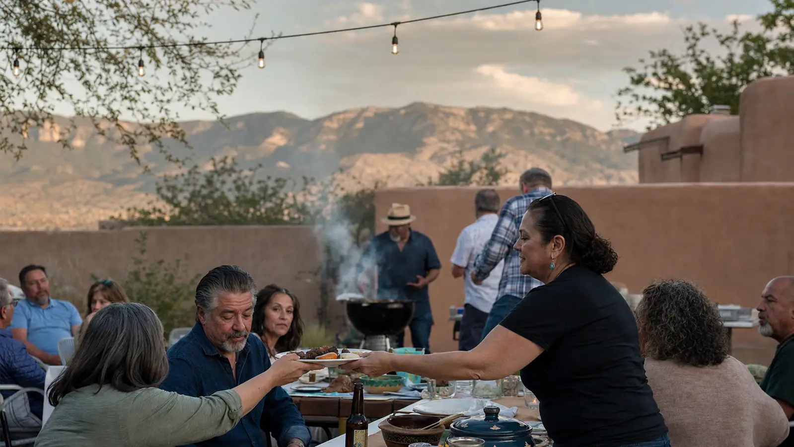 A backyard graduation gathering with folding tables, string lights, and a smoking kettle grill in the early evening