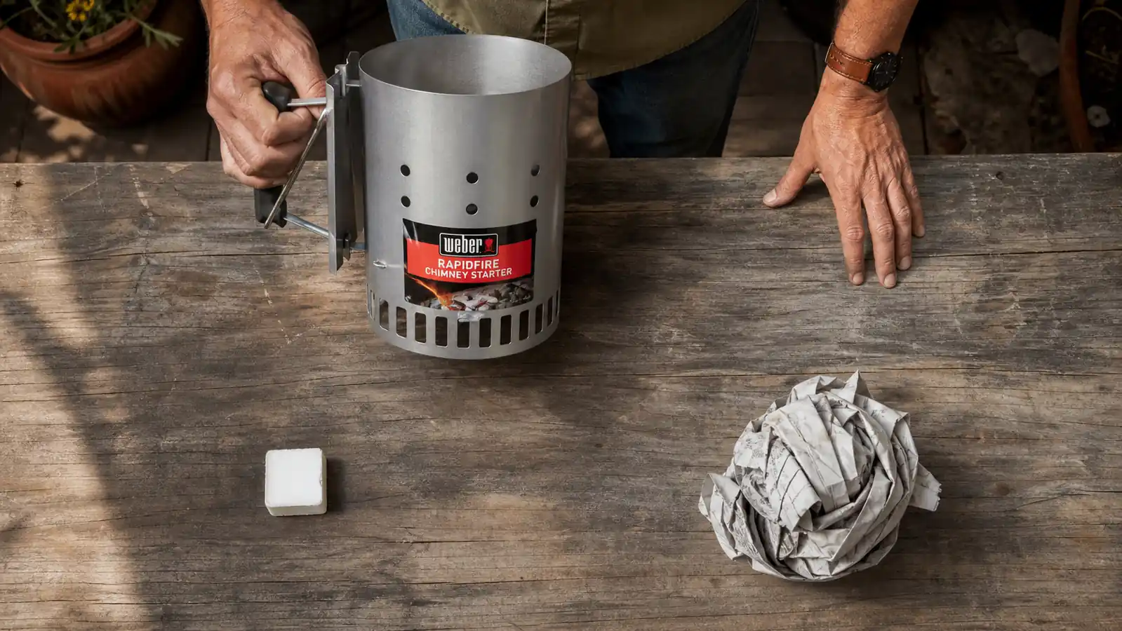A man's hand comparing a compressed paraffin starter cube against a wad of crumpled newspaper on a workbench