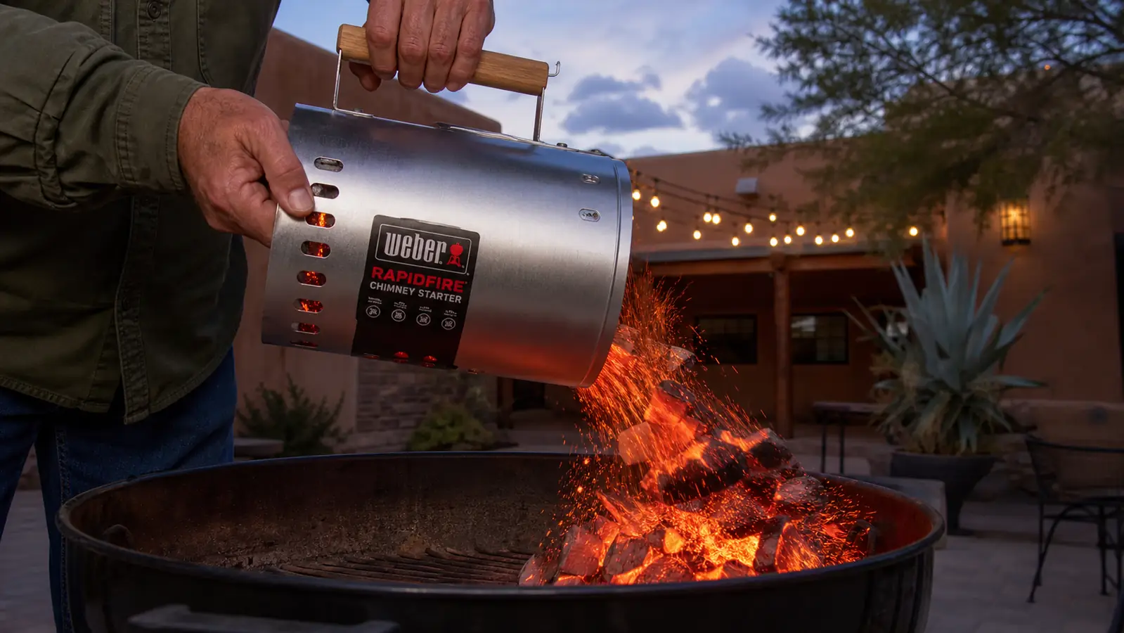 A man's hand gripping the wood handle of a chimney starter and tipping glowing charcoal into an open kettle grill