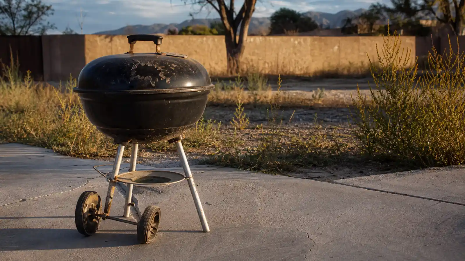 A weathered budget kettle grill sitting in a driveway with a cracked wheel axle and bubbled paint on the lid, no people